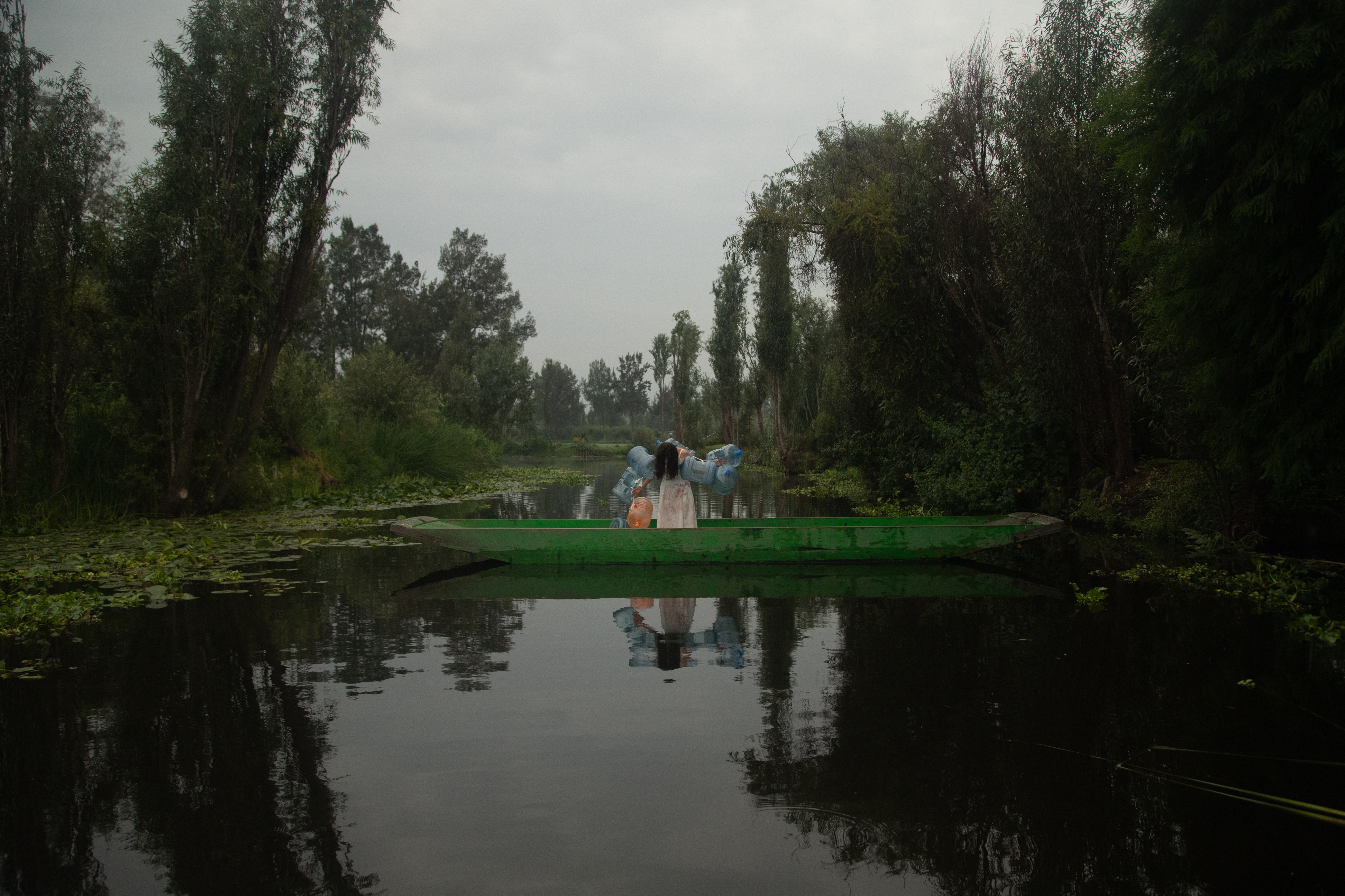 A dark green river flanked by lush green trees with lily pads floating atop the water. In the centre of the river, a long green canoe, a person stands inside, their shoulders adorned with six large blue plastic water containers. They hold an orange plastic water container in their right hand. Their head sags and their long black hair covers their face.